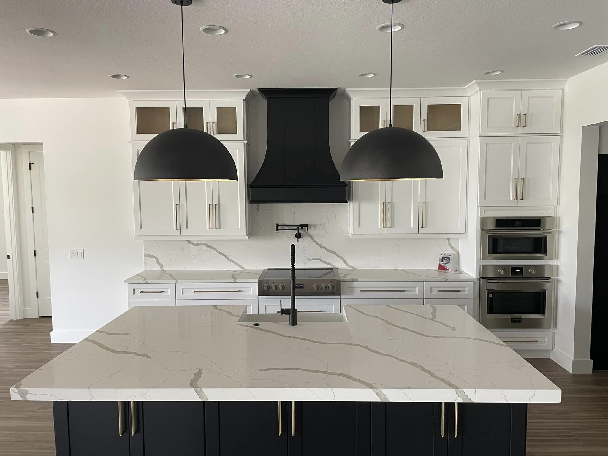 A broad veined island with dark cabinetry, black pendants, and a light kitchen beyond